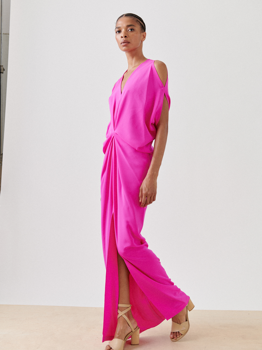 A person wears the Long Miu Dress by Zero + Maria Cornejo, featuring bright pink silk, cut-out shoulders, and a thigh-high slit. They stand against a simple light background with neutral makeup and neatly styled hair.