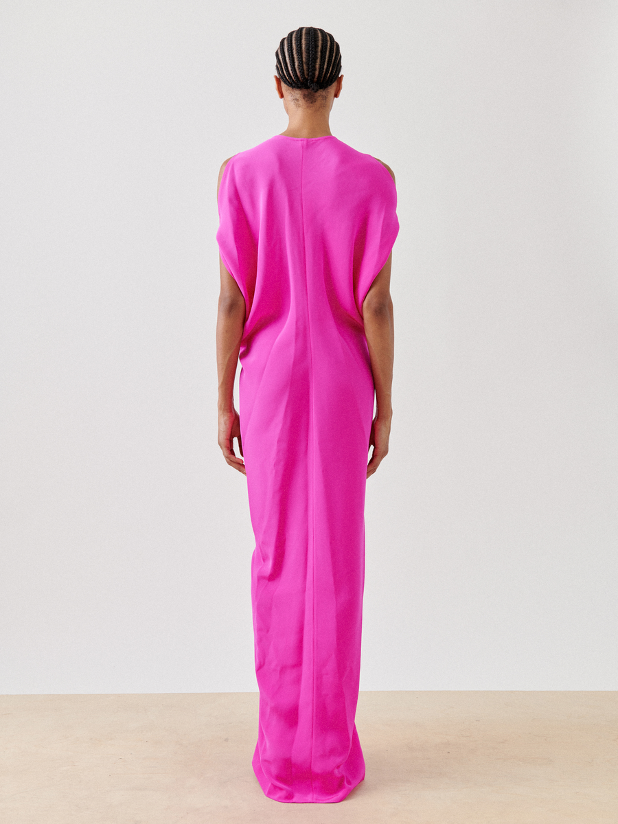 With their hair styled in cornrows, a person stands against a plain wall wearing the Long Miu Dress by Zero + Maria Cornejo. The bright pink, floor-length dress features a sleeveless design with cut-out shoulders and offers a loose fit.