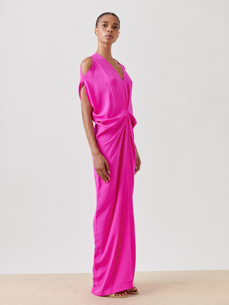 Wearing the Long Miu Dress by Zero + Maria Cornejo, a person stands in vibrant pink silk against a plain light background. This stunning dress features elegant cut-out shoulders and a gathered waist, showcasing exquisite craftsmanship.