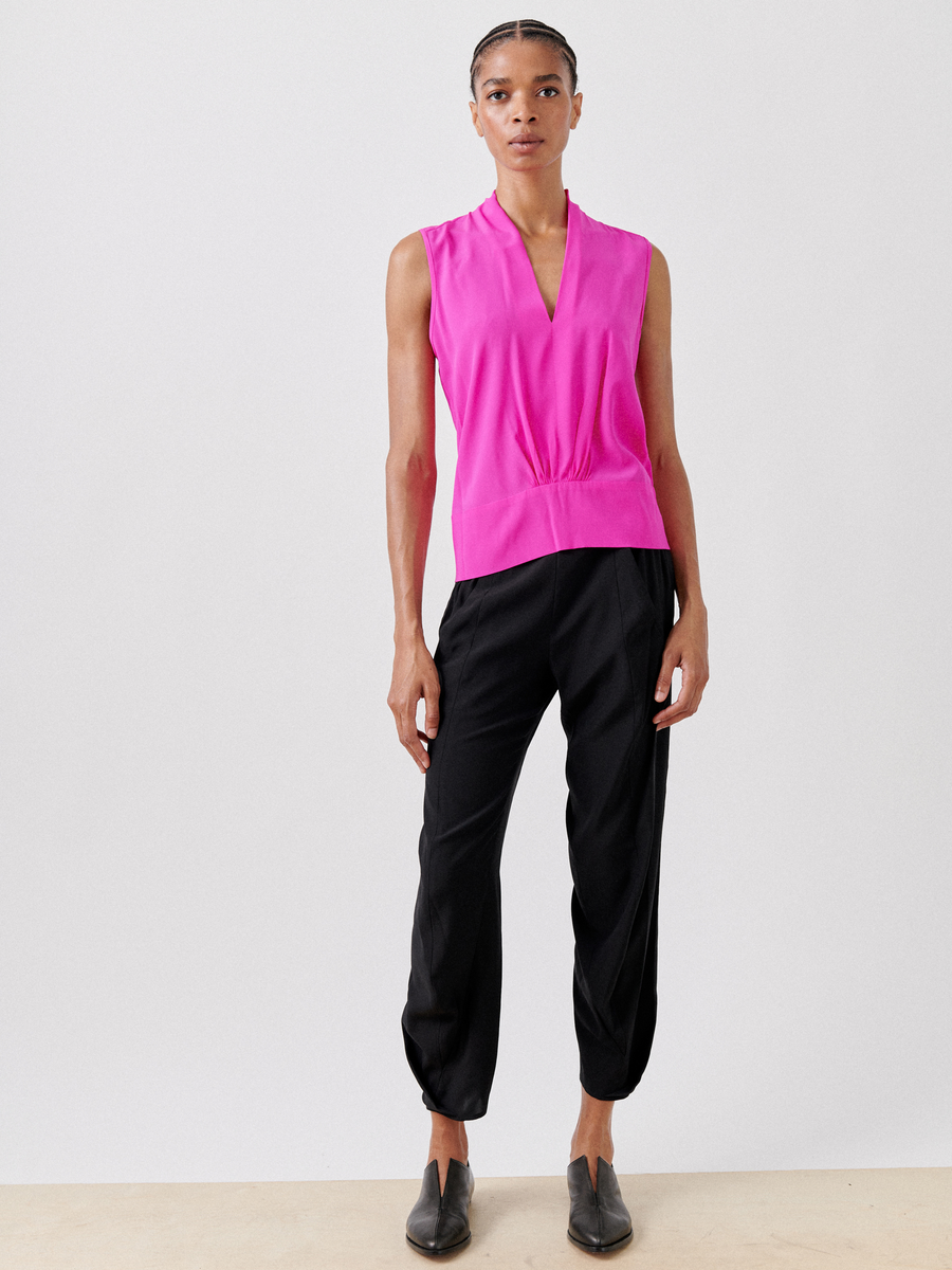 A person poses against a plain white background, wearing the Sacha Top by Zero + Maria Cornejo, featuring a sleeveless v-neckline in bright pink natural fiber, paired with loose black pants and black slip-on shoes. They have short braided hair, a neutral expression, and hands by their sides.