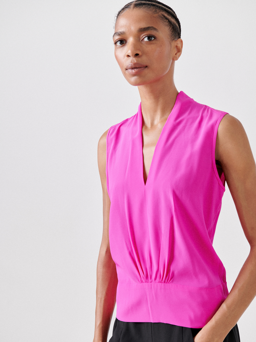 A person wearing the Sacha Top by Zero + Maria Cornejo—a bright pink silk charmeuse sleeveless blouse with a deep V-neck and gathered waist—and black pants stands against a light gray background with a neutral expression.