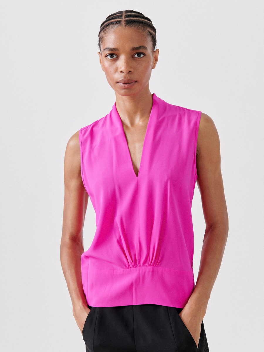 Wearing the vibrant Sacha Top by Zero + Maria Cornejo, a person with short braided hair stands with hands in black pants' pockets. The sleeveless pink charmeuse top features a V-neck and loose fit as they gaze directly at the camera against a white background.