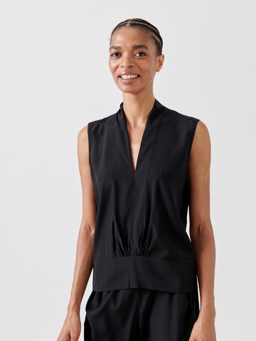 A smiling person with braided hair is wearing a sleeveless black Sacha Top by Zero + Maria Cornejo and black pants. The Sacha Top, made in New York, features a deep V-neckline and a gathered waist detail. The background is plain white.