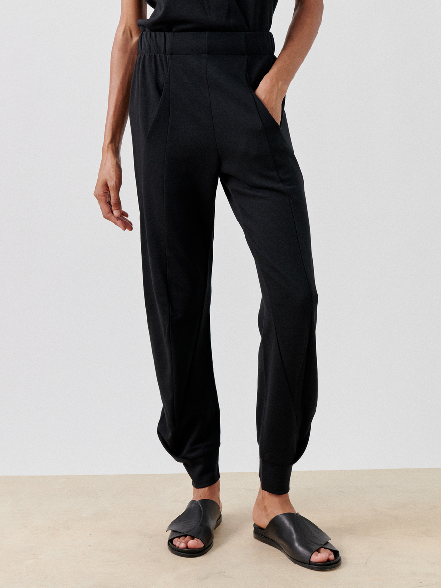 A person wearing black Jersey Cuff Akeo Pants by Zero + Maria Cornejo with an elastic banded waist and black open-toe slip-on sandals stands against a plain backdrop. One hand is in a hidden front pocket while the other rests by their side. The outfit, made from cotton-modal jersey fabric, emphasizes comfort and casual style.