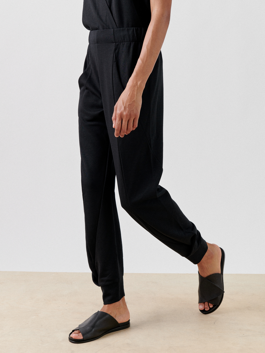 A person is partially shown from the neck down, wearing Jersey Cuff Akeo Pants by Zero + Maria Cornejo featuring an elastic banded waist and a black shirt. They are walking on a beige surface, and their feet are visible in black slide sandals. The background is plain white.