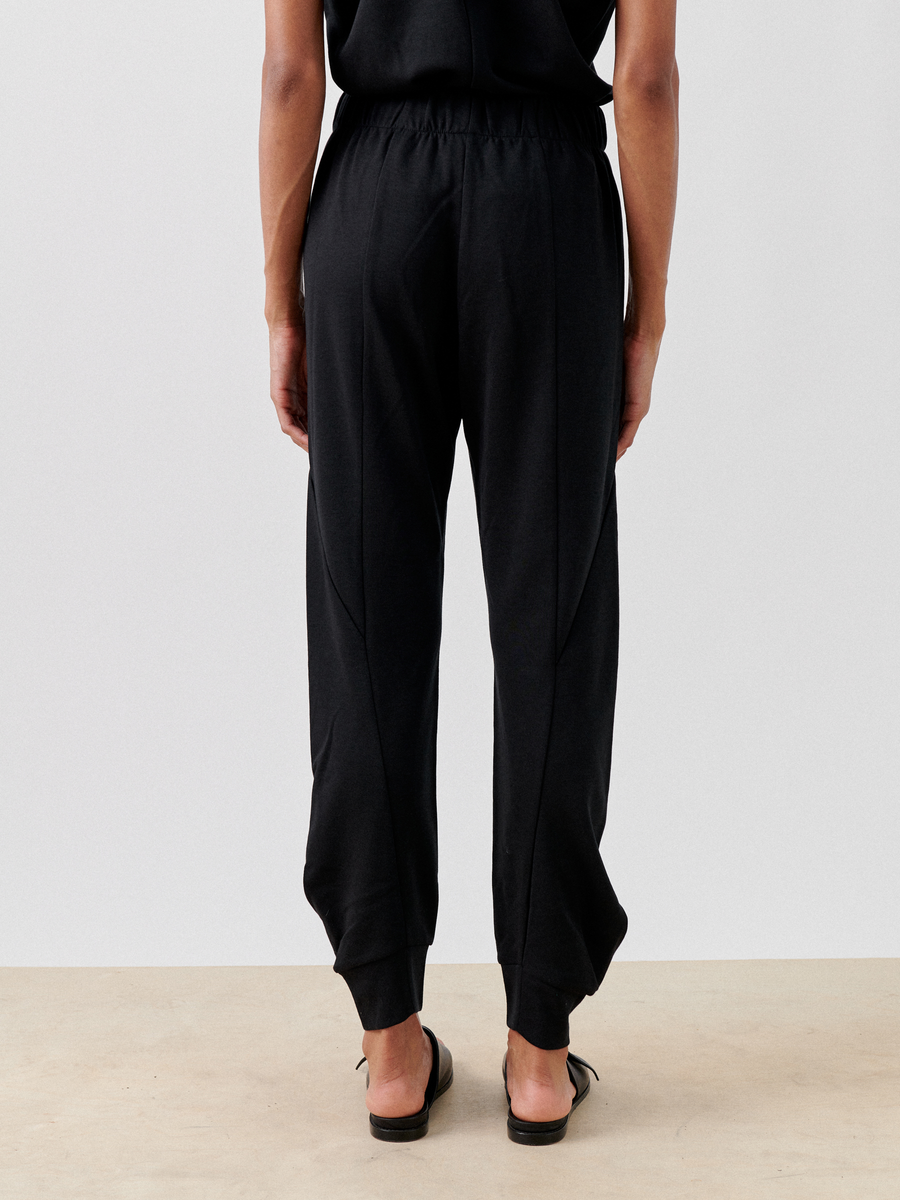 A person stands facing away from the camera, wearing Zero + Maria Cornejo Jersey Cuff Akeo Pant, black cotton-modal jogger pants with an elastic banded waist and cuffs at the ankles. They are also wearing black flat sandals. The background is plain white.