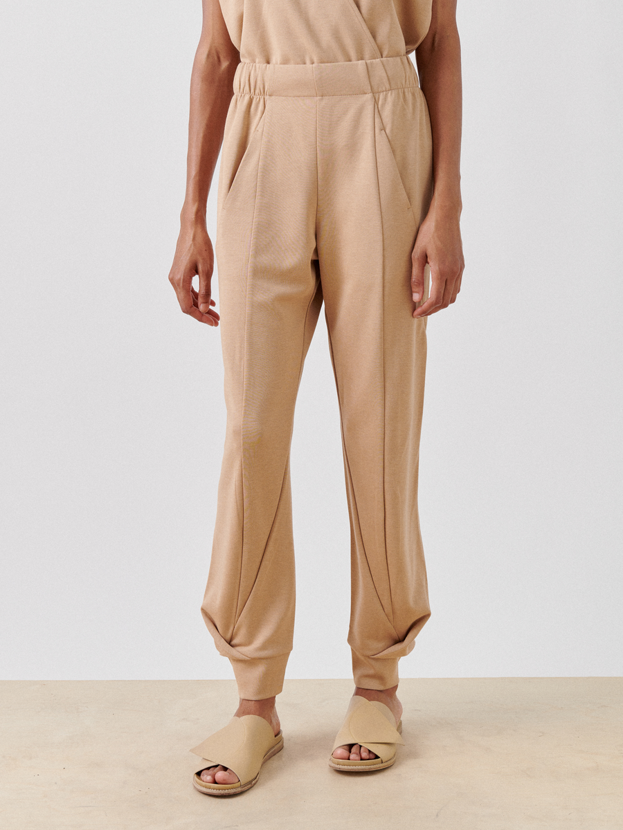 A person stands against a plain background, wearing the Zero + Maria Cornejo Jersey Cuff Akeo Pant with pin-tuck detailing and hidden front pockets. The cotton-modal jersey pants have an elastic banded waist, offering a relaxed fit that tapers at the ankles. Tan open-toe slip-on sandals complete the look as their arms rest at their sides.