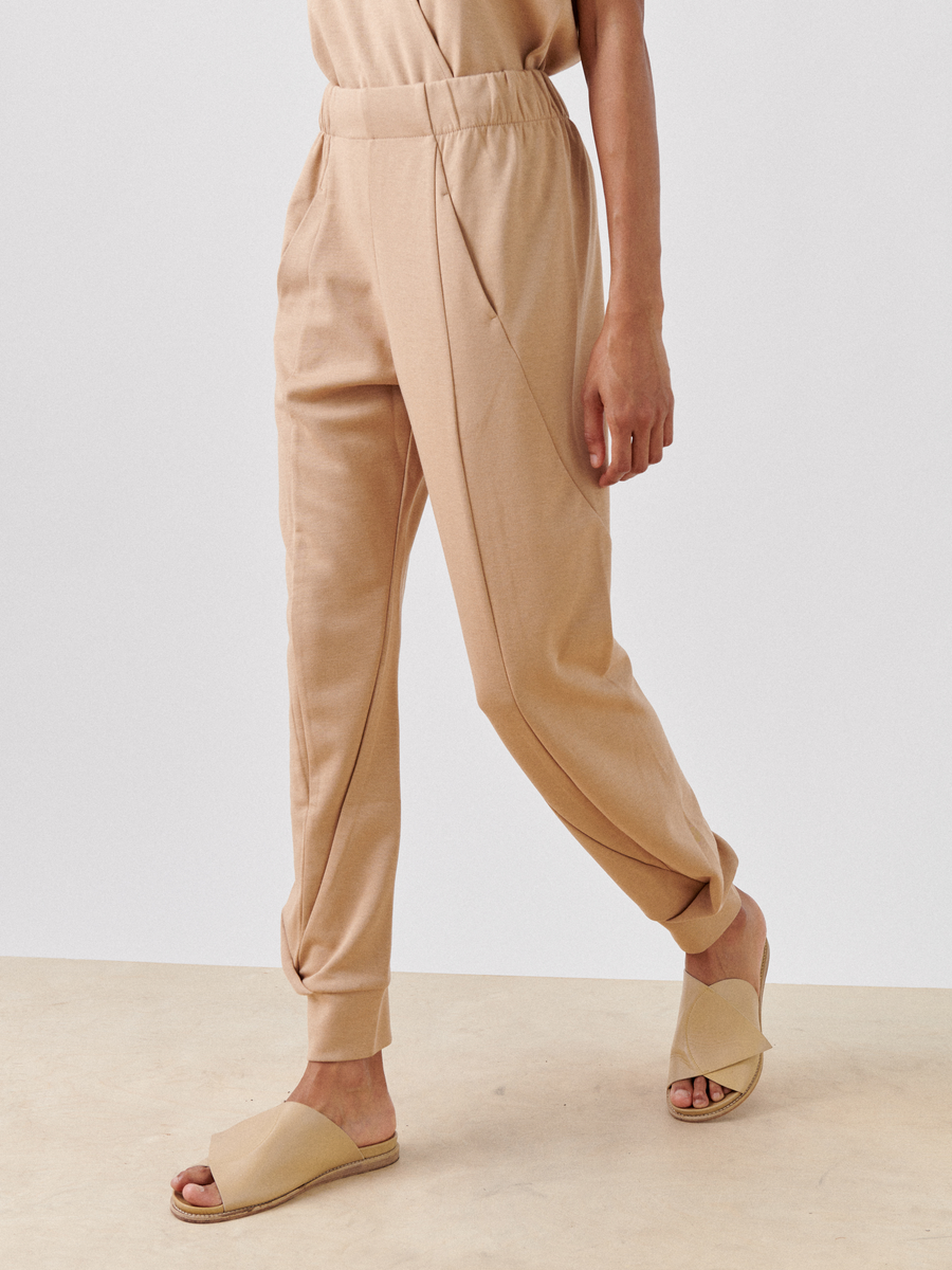 A person, wearing Zero + Maria Cornejo Jersey Cuff Akeo Pant with an elastic-banded waist and hidden front pockets, walks on a light-colored floor. The relaxed-fit pants are paired with beige sandals featuring an open-toe design and wide straps. The image focuses on the lower body, leaving the upper body out of view.