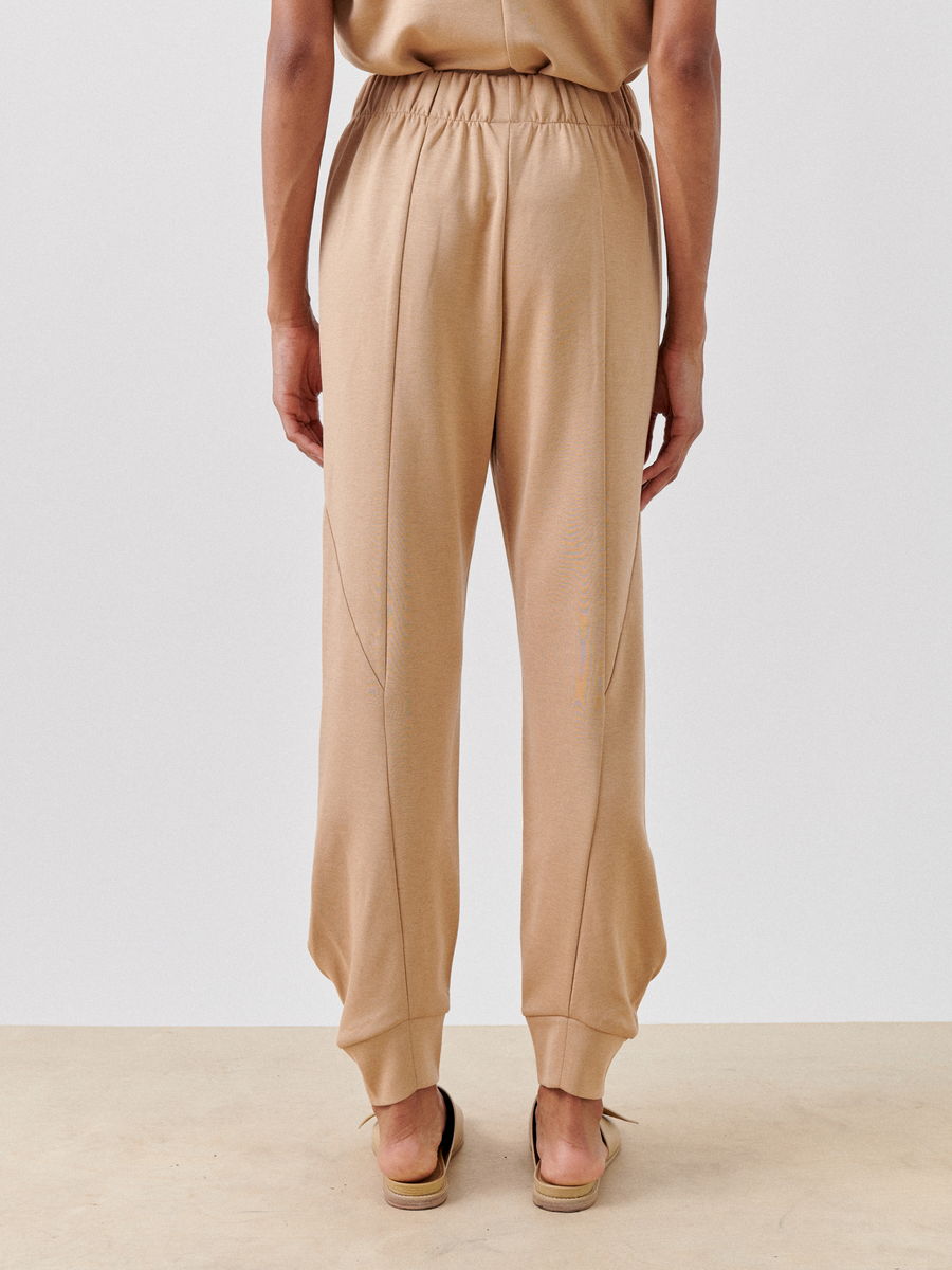Back view of a person wearing beige jogger pants and sandals. The Jersey Cuff Akeo Pant by Zero + Maria Cornejo have an elastic banded waist and ankle cuffs, with seam line details running down the back of the legs. The person is standing on a light-colored surface against a neutral background.