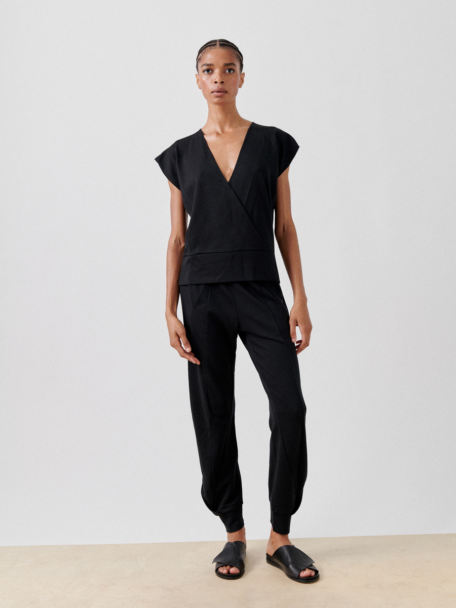 A person stands against a plain backdrop wearing a sleek, black Wrap Mido Top by Zero + Maria Cornejo with matching black pants. They have neatly styled hair and are wearing black open-toed sandals. The overall look is minimalist and modern.