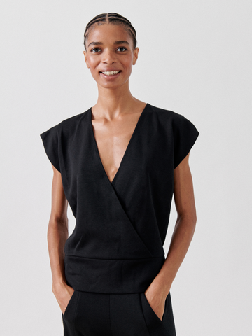 A person with short braided hair smiles at the camera while standing with hands in pockets. They are wearing a sleeveless black Wrap Mido Top by Zero + Maria Cornejo made from soft cotton-modal jersey and matching black pants, set against a plain white background.