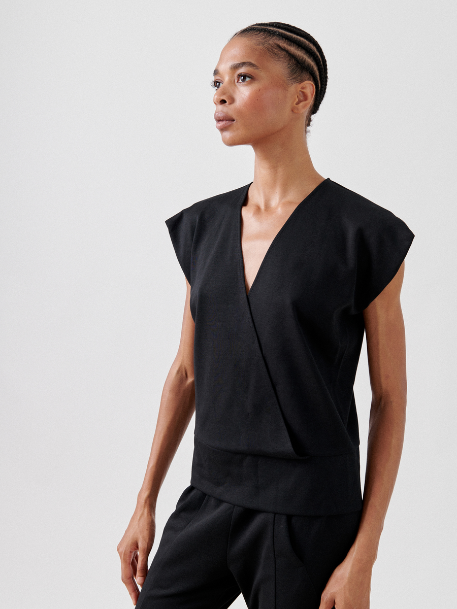 A person with a short braided hairstyle wears a black Wrap Mido Top by Zero + Maria Cornejo with cap sleeves and matching black pants. They stand against a plain light background, looking slightly to the side with a confident expression.
