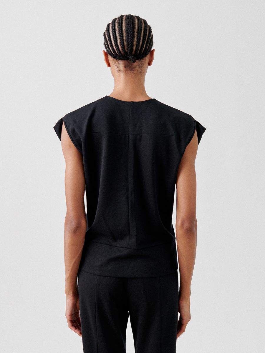 A person with cornrow braids is shown from behind, wearing a sleeveless black *Wrap Mido Top* by *Zero + Maria Cornejo* and black pants made of cotton-modal jersey. The image is set against a plain white background.