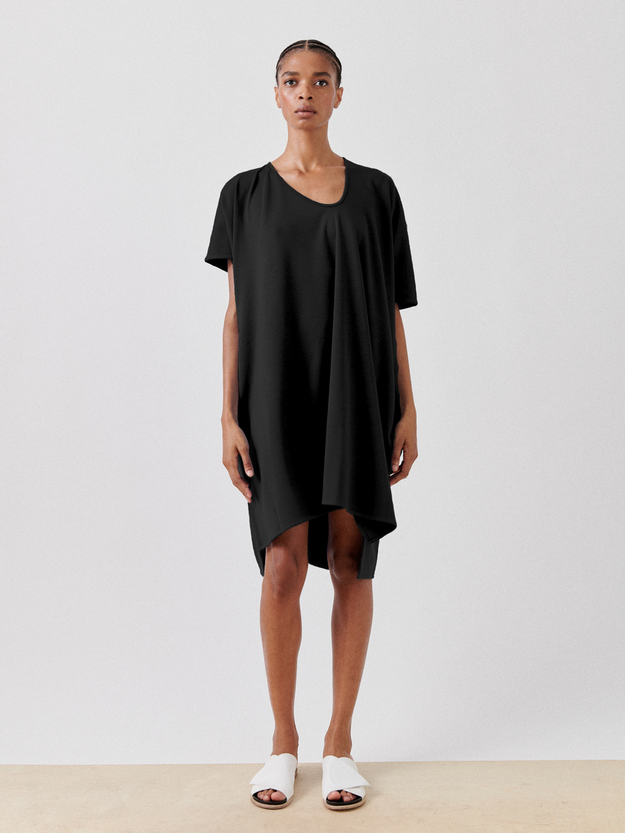A person stands against a plain background, wearing a minimalist and elegant knee-length Zero + Maria Cornejo Jersey Off Kilter Dress with short sleeves and an asymmetrical hem. They have braided hair and are donning white open-toe shoes, epitomizing sustainable fashion.