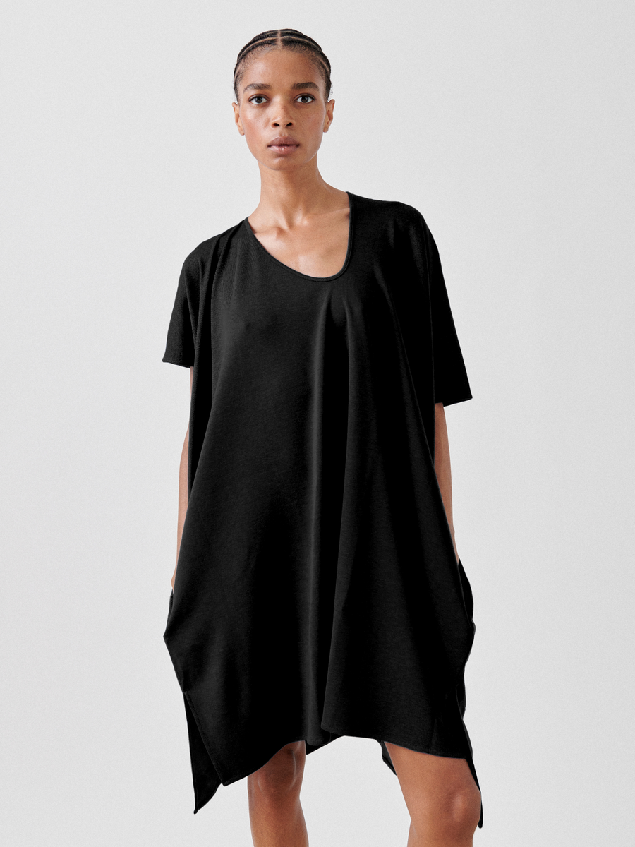 A person stands against a plain background wearing the Jersey Off Kilter Dress by Zero + Maria Cornejo. Made from cotton-modal jersey, the dress features an asymmetrical hemline that falls to the knees. The person has short braided hair and a serious expression, embodying sustainable fashion.