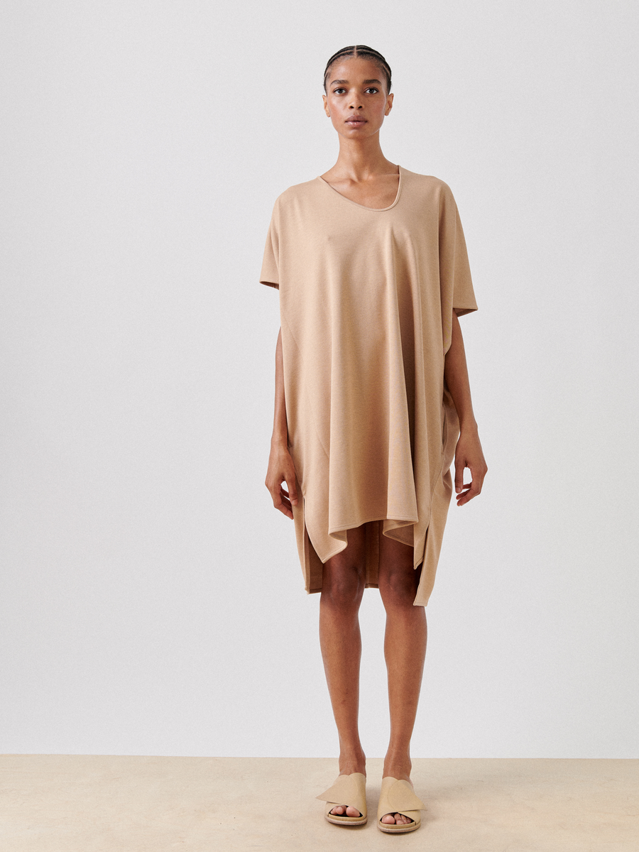 A person stands against a plain background, wearing a loose, knee-length Jersey Off Kilter Dress by Zero + Maria Cornejo with an asymmetrical hemline. They are also wearing beige slide sandals, embodying sustainable fashion. The individual has a neutral facial expression and their hair is styled neatly.