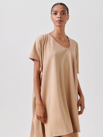 A person is standing against a plain background, wearing a loose-fitting, knee-length Jersey Off Kilter Dress from Zero + Maria Cornejo made from cotton-modal jersey. They have short, styled hair and a neutral expression, with one hand resting by their side and the other slightly raised.
