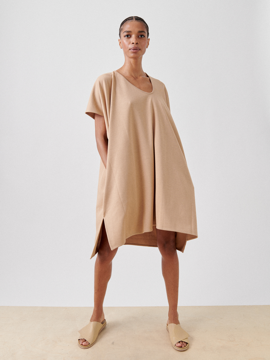 A person stands against a plain background, wearing the Jersey Off Kilter Dress by Zero + Maria Cornejo. It is a loose-fitting, knee-length dress made from sustainable cotton-modal jersey with short sleeves and side slits. They have their hands behind their back and are also wearing tan slide sandals. Their hair is styled in neat, tight braids.