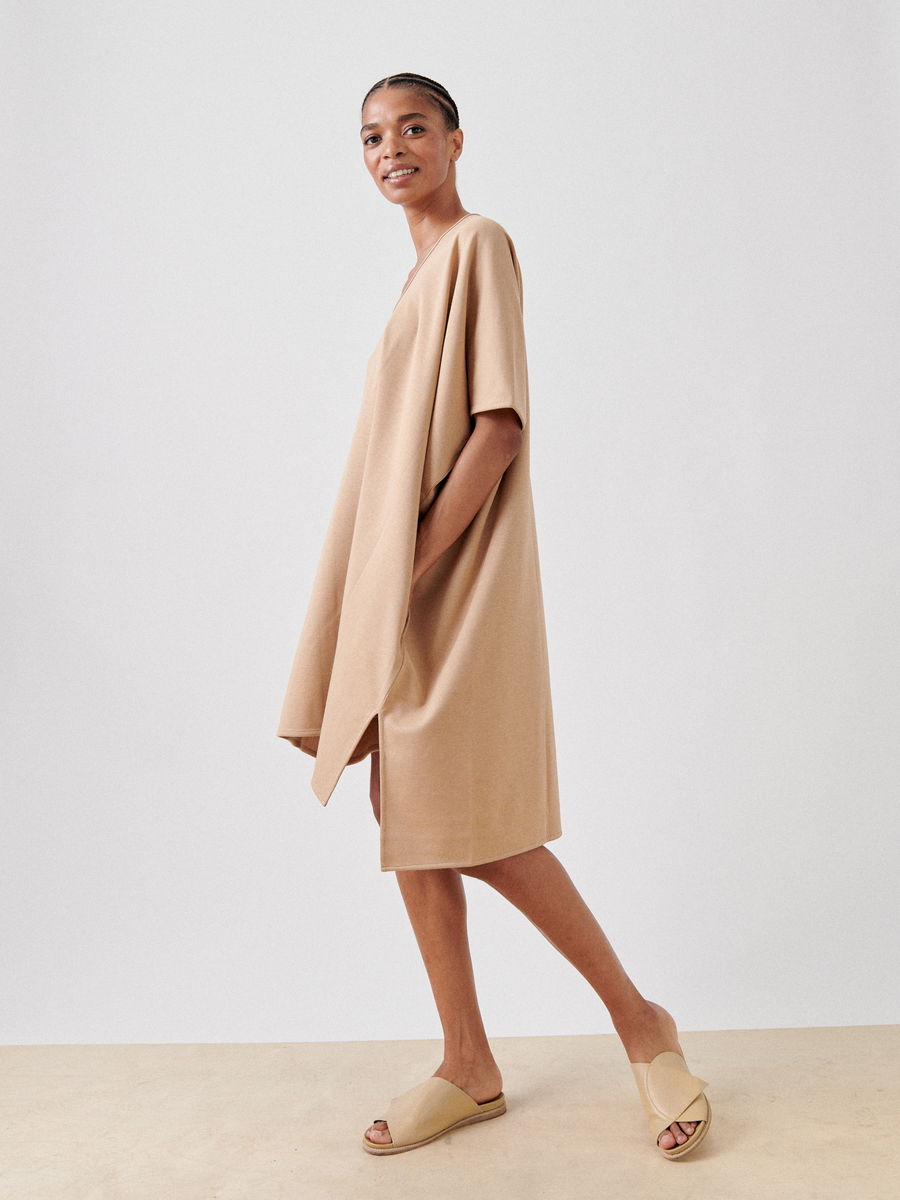 A person stands sideways, smiling, wearing the Jersey Off Kilter Dress by Zero + Maria Cornejo with short sleeves and beige slip-on sandals. The background is plain white, and the person's hair is pulled back. This outfit highlights the ease of sustainable fashion with its comfortable cotton-modal jersey fabric.