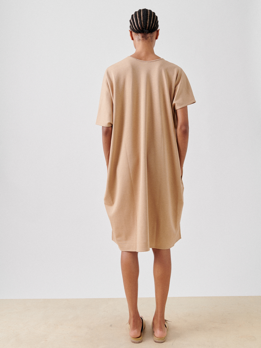 A person with braided hair is standing with their back to the camera, wearing a loose-fitting, knee-length, beige Jersey Off Kilter Dress by Zero + Maria Cornejo. They are also wearing tan slip-on shoes. The background is plain white, highlighting an image of sustainable fashion.