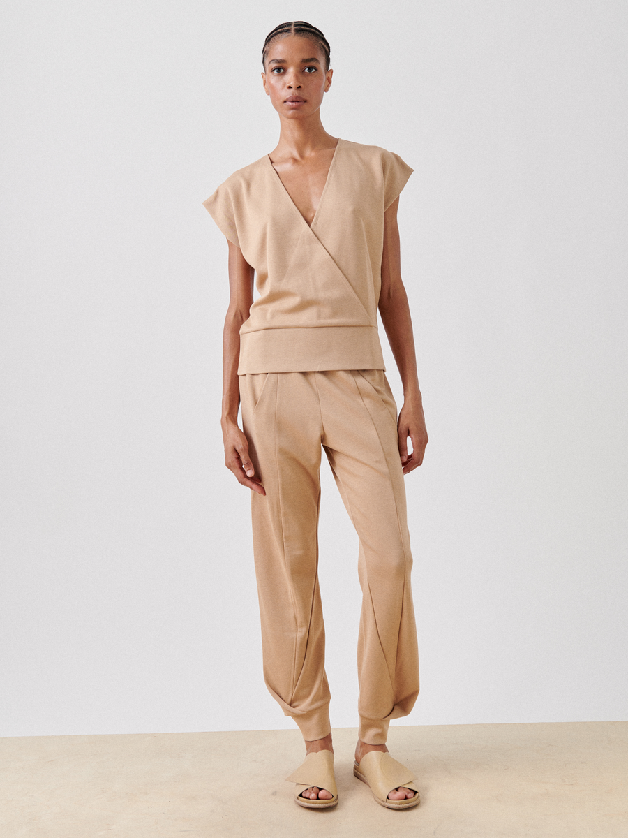 A person stands against a plain background wearing a beige, Wrap Mido Top from Zero + Maria Cornejo, paired with matching loose-fitting pants that taper at the ankles. They are also wearing beige sandals, and their hair is braided and pulled back.