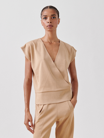 A person stands against a plain white background, wearing a tan Wrap Mido Top by Zero + Maria Cornejo with short, slightly flared sleeves, and matching tan pants. The person's hair is styled in tight braids, and they have a neutral expression, with one hand in their pocket.