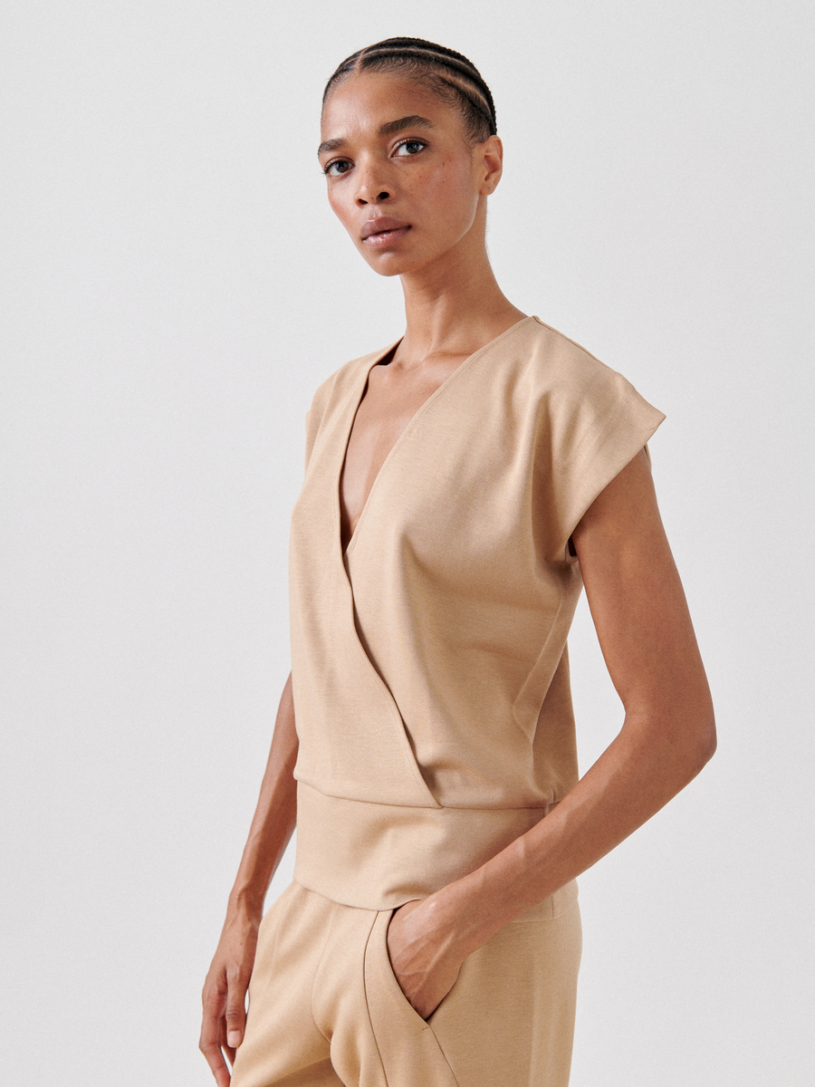 A person with braided hair poses confidently in a minimalist, beige outfit. The ensemble features the Wrap Mido Top from Zero + Maria Cornejo and matching trousers made from comfortable cotton-modal jersey. The plain background enhances the focus on the individual and their stylish clothing.