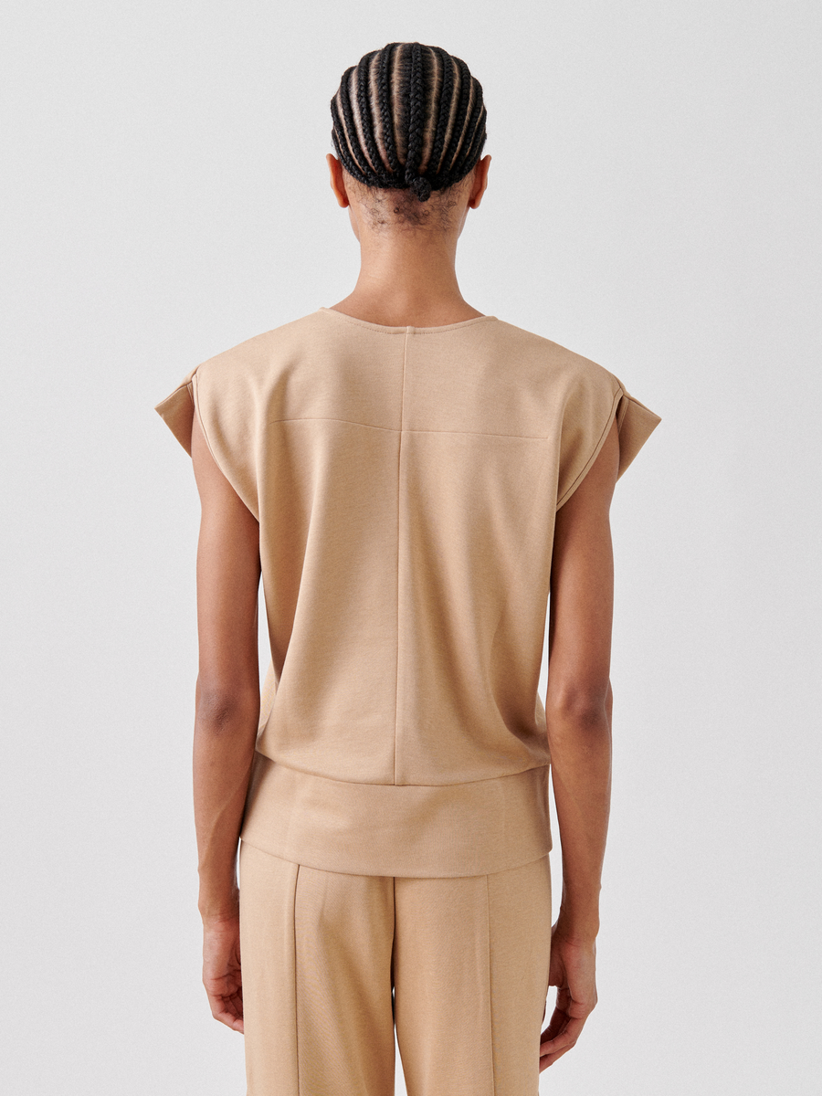 A person stands facing away from the camera, showcasing a beige, sleeveless Wrap Mido Top by Zero + Maria Cornejo paired with beige trousers. The cotton-modal jersey top features a simple, clean design with a straight hem. They have neatly braided hair and are set against a light gray background.