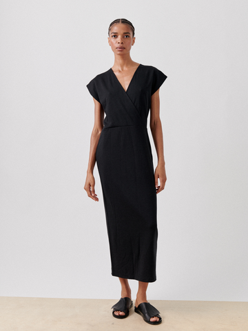 A person with a calm expression stands against a plain background, wearing the Wrap Mido Dress by Zero + Maria Cornejo, which is a sleeveless black v-neck faux wrap dress that falls below the knees. They are also wearing black slide sandals. Their hair is styled in a tidy manner, and they have a composed posture.