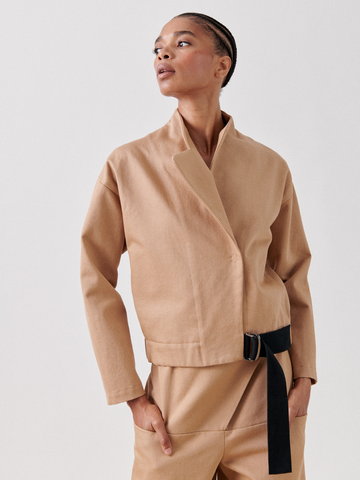 A person poses confidently in a minimalist fashion style. They are wearing the Zero + Maria Cornejo Long-Sleeved Edi Bomber with an adjustable grosgrain belt, paired with matching tan pants. The background is plain and light-colored, emphasizing the clean, modern look of the outfit.