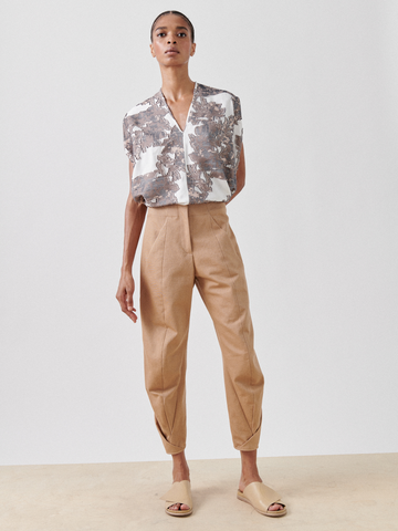 A person stands against a plain background, wearing a short-sleeved, button-up shirt with a geometric pattern, tucked into high-waisted beige Takeo Pant from Zero + Maria Cornejo made from GOTS-certified cotton. They are also wearing beige sandals and pose with a neutral expression, hands by their sides.