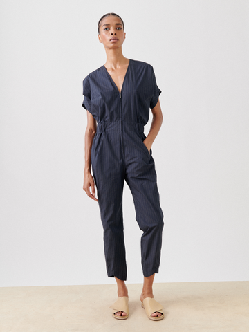 A person stands against a plain white background, wearing the Aissa V-Neck Jumpsuit by Zero + Maria Cornejo. The dark blue pinstriped jumpsuit is crafted from recycled textile waste and features short sleeves, an elastic waistband, and ankle-length pants. The person is also wearing beige slip-on shoes and has their hair pulled back.
