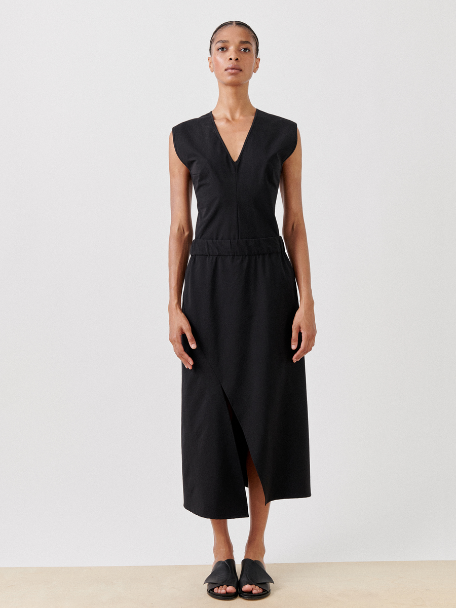 A person with a neutral expression is standing against a plain background. They are wearing a sleeveless, V-neck black dress with a waist seam and a wrap-style front made of eco-hybrid fabric. They are also wearing the Bias Slip Skirt by Zero + Maria Cornejo and black sandals. The overall look is minimalistic and elegant.