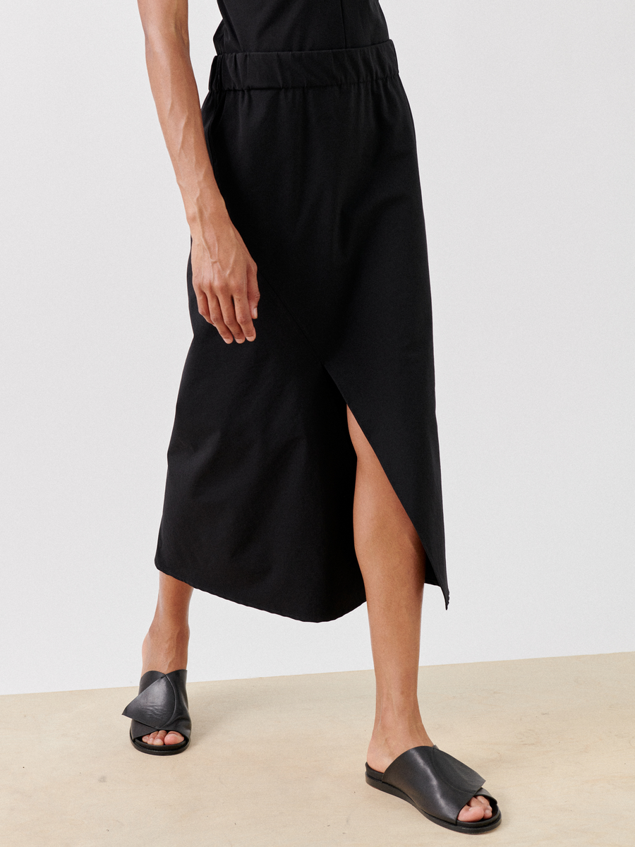 Person wearing a black, asymmetrical, eco-hybrid fabric Bias Slip Skirt from Zero + Maria Cornejo that extends to mid-calf and black open-toe footwear with a unique strap design. The photo captures the lower half of the figure standing on a beige surface with a white background.