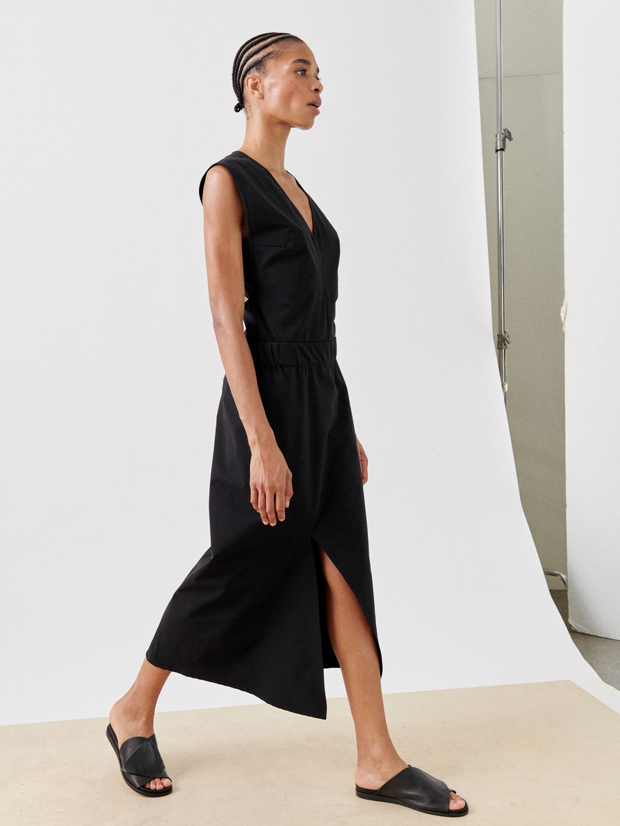 A person wearing a sleeveless black wrap dress and black slide sandals is walking against a plain, light-colored background. They have short braided hair and are captured in profile. The dress, featuring a deep V-neck and a front slit, flows seamlessly like an elegant Bias Slip Skirt by Zero + Maria Cornejo.