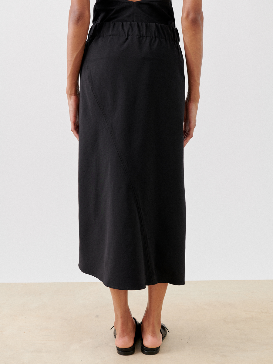The image shows a person standing with their back to the camera, wearing a black Bias Slip Skirt from Zero + Maria Cornejo made from an eco-hybrid fabric. The skirt has a high waist and an asymmetrical seam running diagonally from the left waist to the right hem. The person is also wearing black flat sandals.