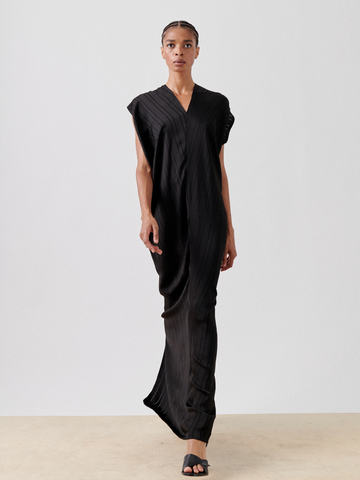 A woman wearing a black, sleeveless, V-neck Long Issa Dress by Zero + Maria Cornejo with a gathered detail and pleated design walks forward. She has braided hair and wears black high-heeled mules. The background is a plain, light-colored wall and floor.