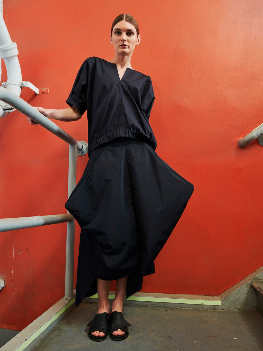 A person stands confidently on a stairwell with an orange wall backdrop, wearing a loose, dark blue outfit featuring the Nio Skirt by Zero + Maria Cornejo and black sandals. Their long brown hair is pulled back into a geometric silhouette.