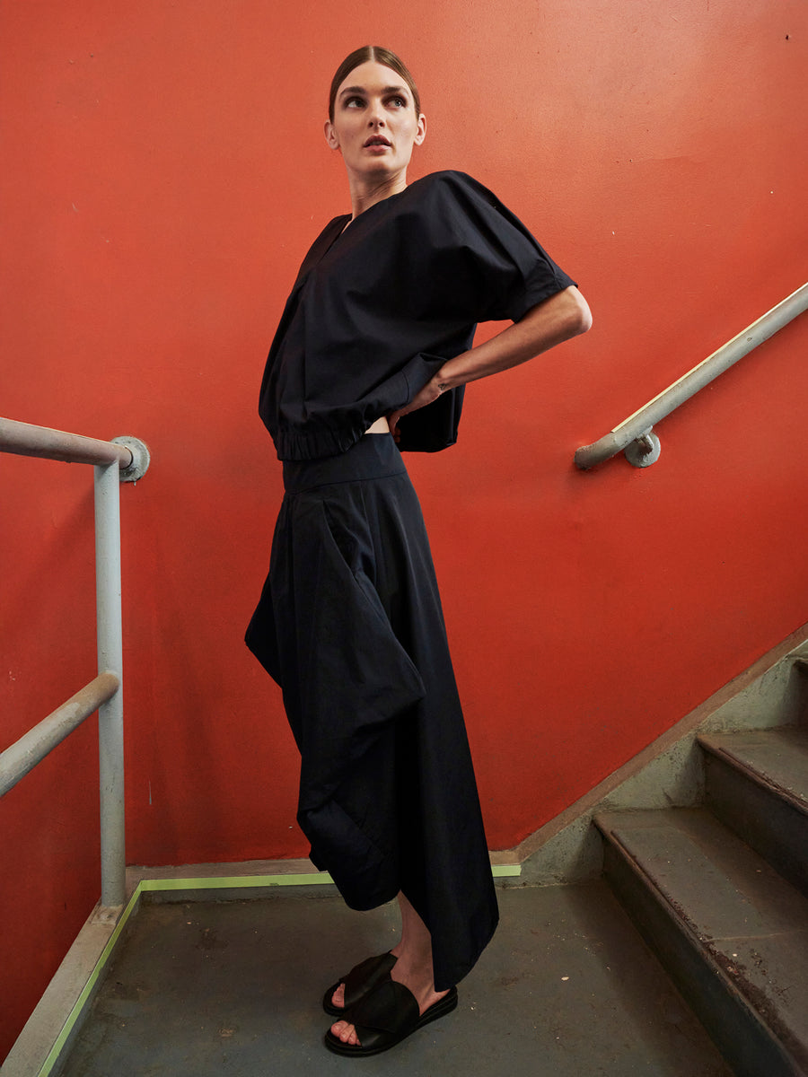 A person confidently poses on stairs against a vibrant orange wall, wearing a stylish black Nio Skirt from Zero + Maria Cornejo. The outfit features loose, flowing pants with a geometric silhouette and black sandals, with their hair neatly tied back and hands resting on their waist.