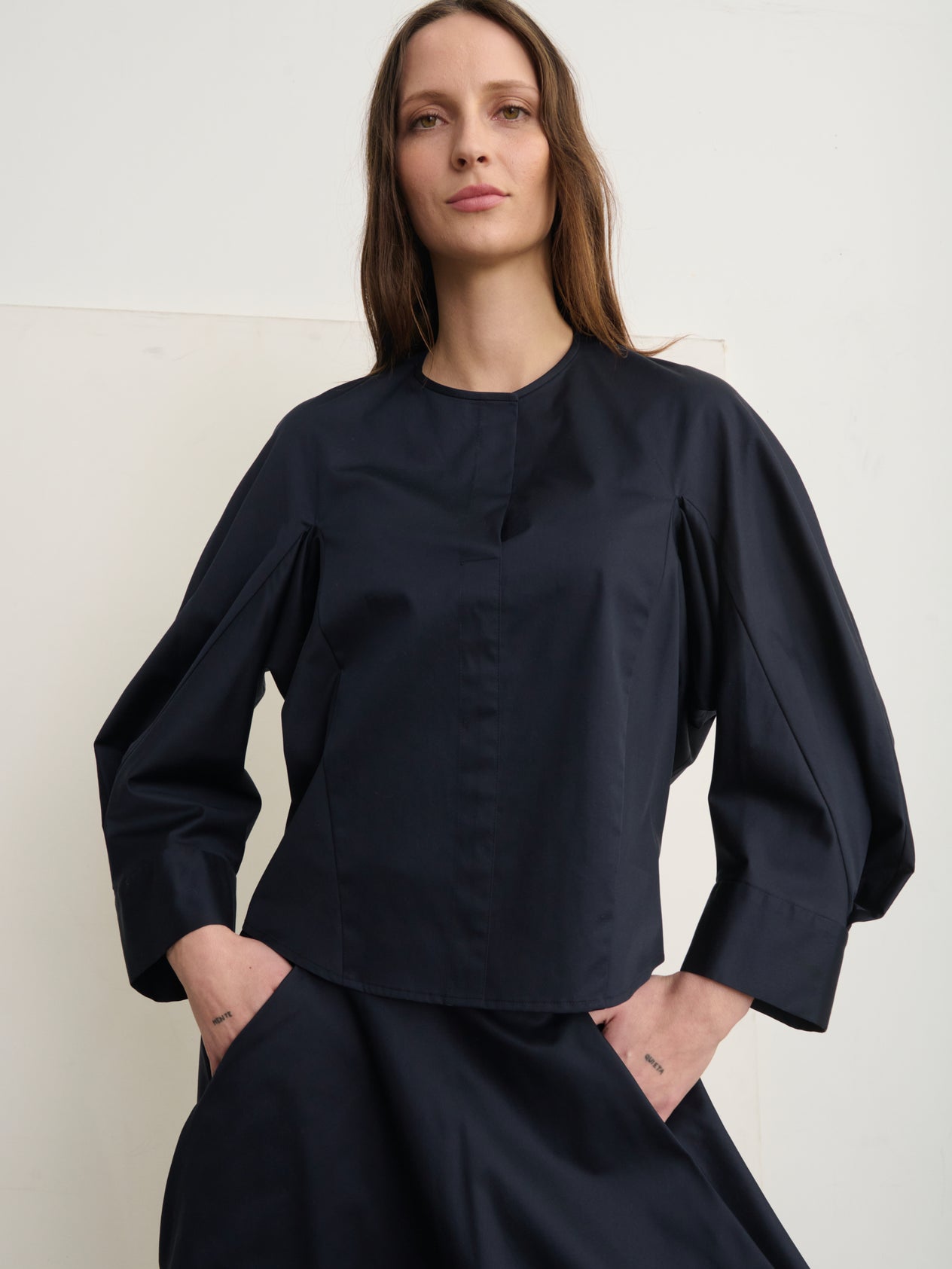 Against a concrete wall, someone stands wearing Zero + Maria Cornejo's Long-Sleeved Akeo Shirt with voluminous sleeves and blue high-waisted pants. The shirt's round neckline complements their composed expression as they place their hands on their hips.