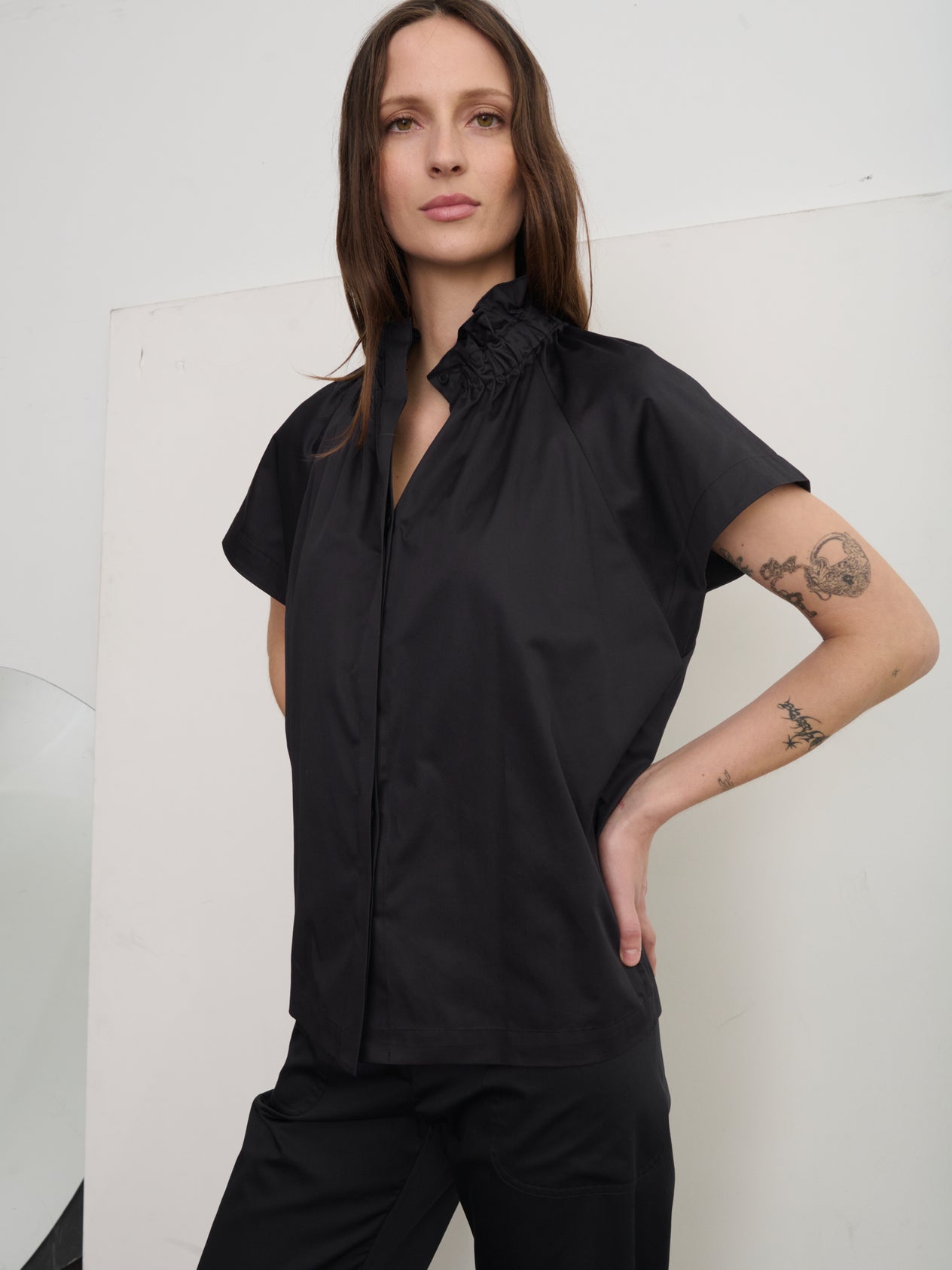 A person with long brown hair stands confidently, hands on hips, in a stylish Elastic Gaban Shirt by Zero + Maria Cornejo, featuring a gathered collar. Tattoos adorn their arm as the neutral-toned wall behind complements their chic ensemble.