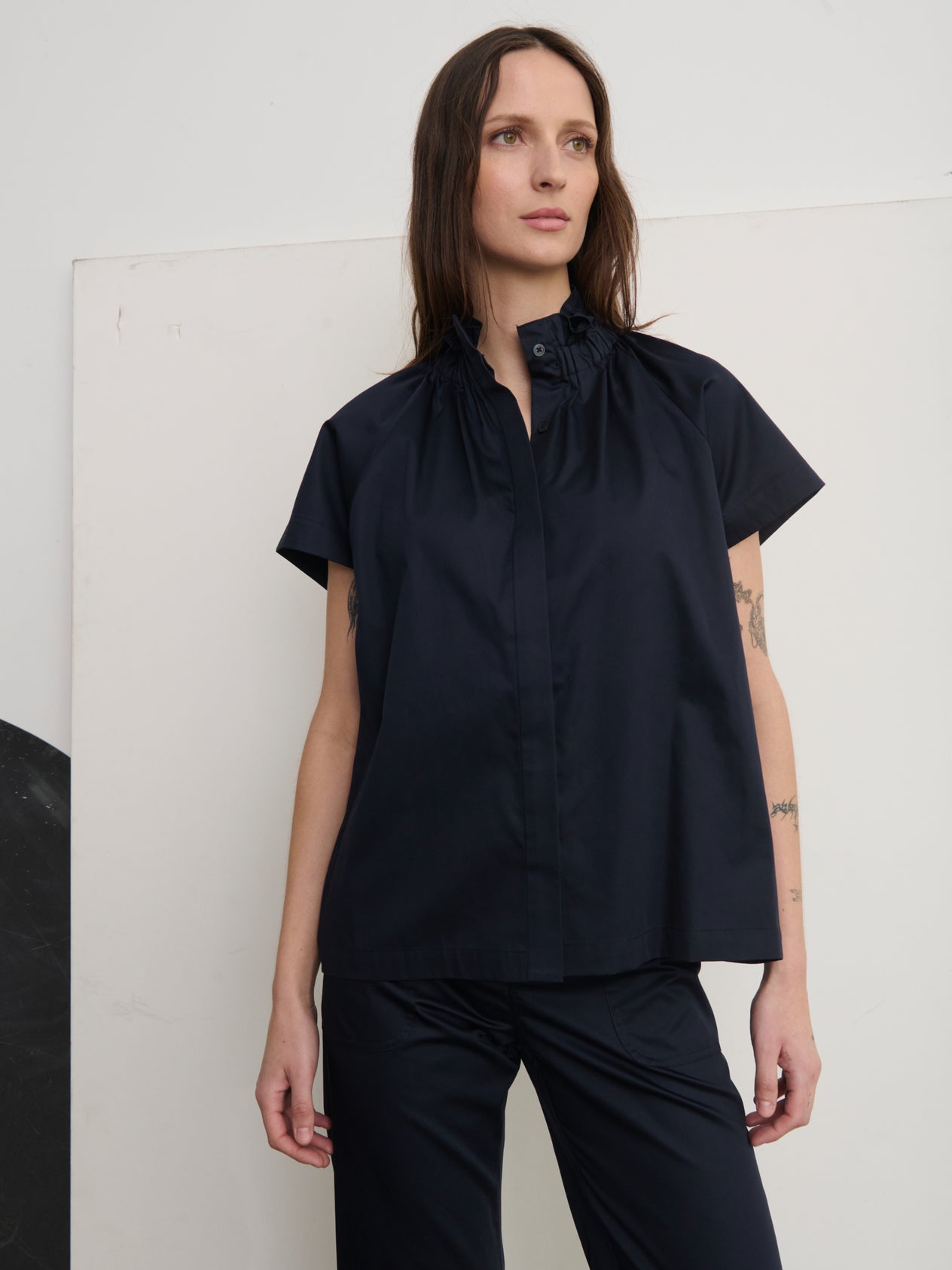 A person with long brown hair stands confidently, hands on hips, in a stylish Elastic Gaban Shirt by Zero + Maria Cornejo, featuring a gathered collar. Tattoos adorn their arm as the neutral-toned wall behind complements their chic ensemble.