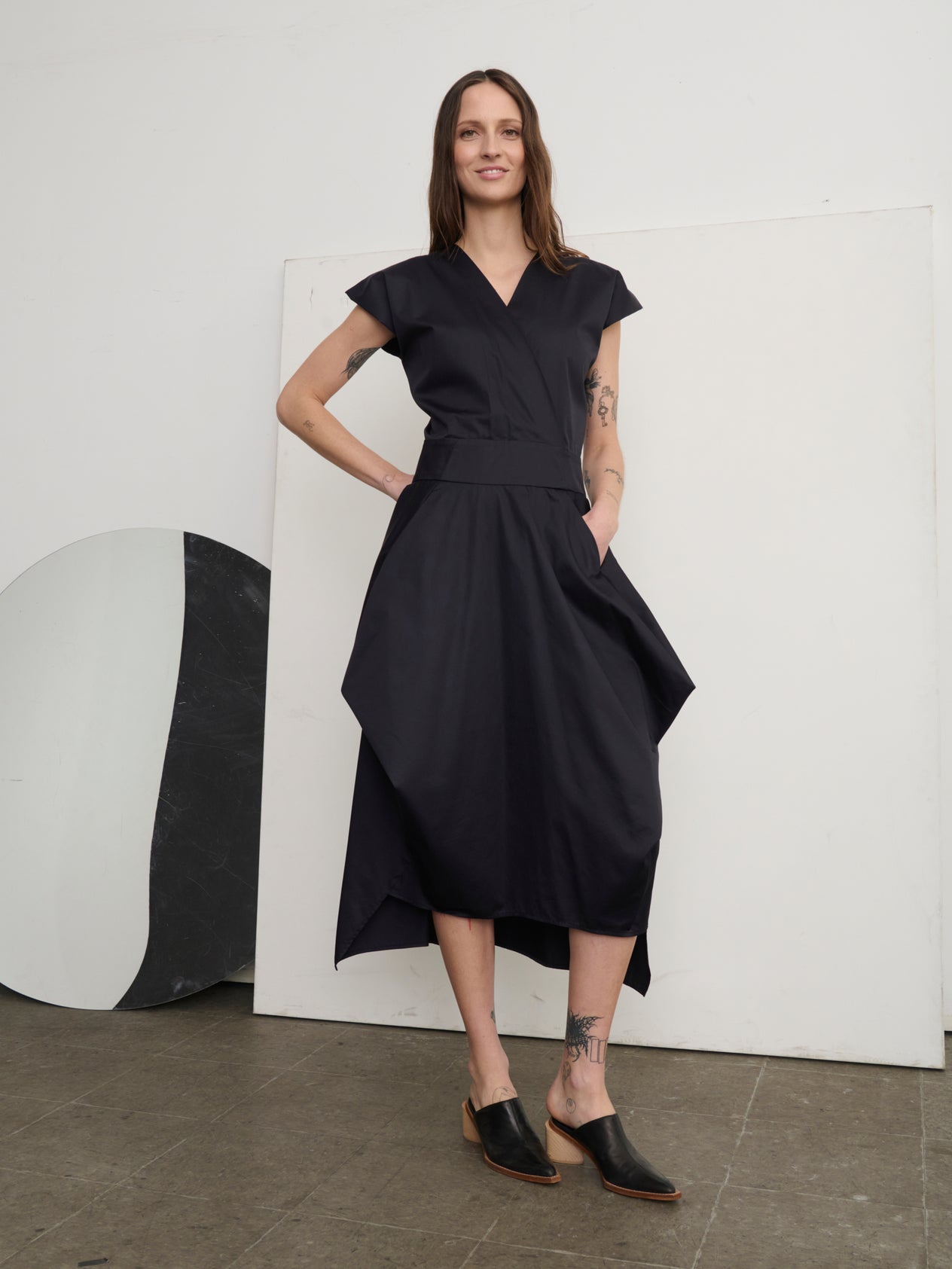 A person with long brown hair confidently wears a sleeveless, dark Wrap Nio Dress by Zero + Maria Cornejo. Tattoos adorn one arm, and they sport black slip-on shoes. Abstract shapes and a neutral wall in the background enhance the geometric charm.