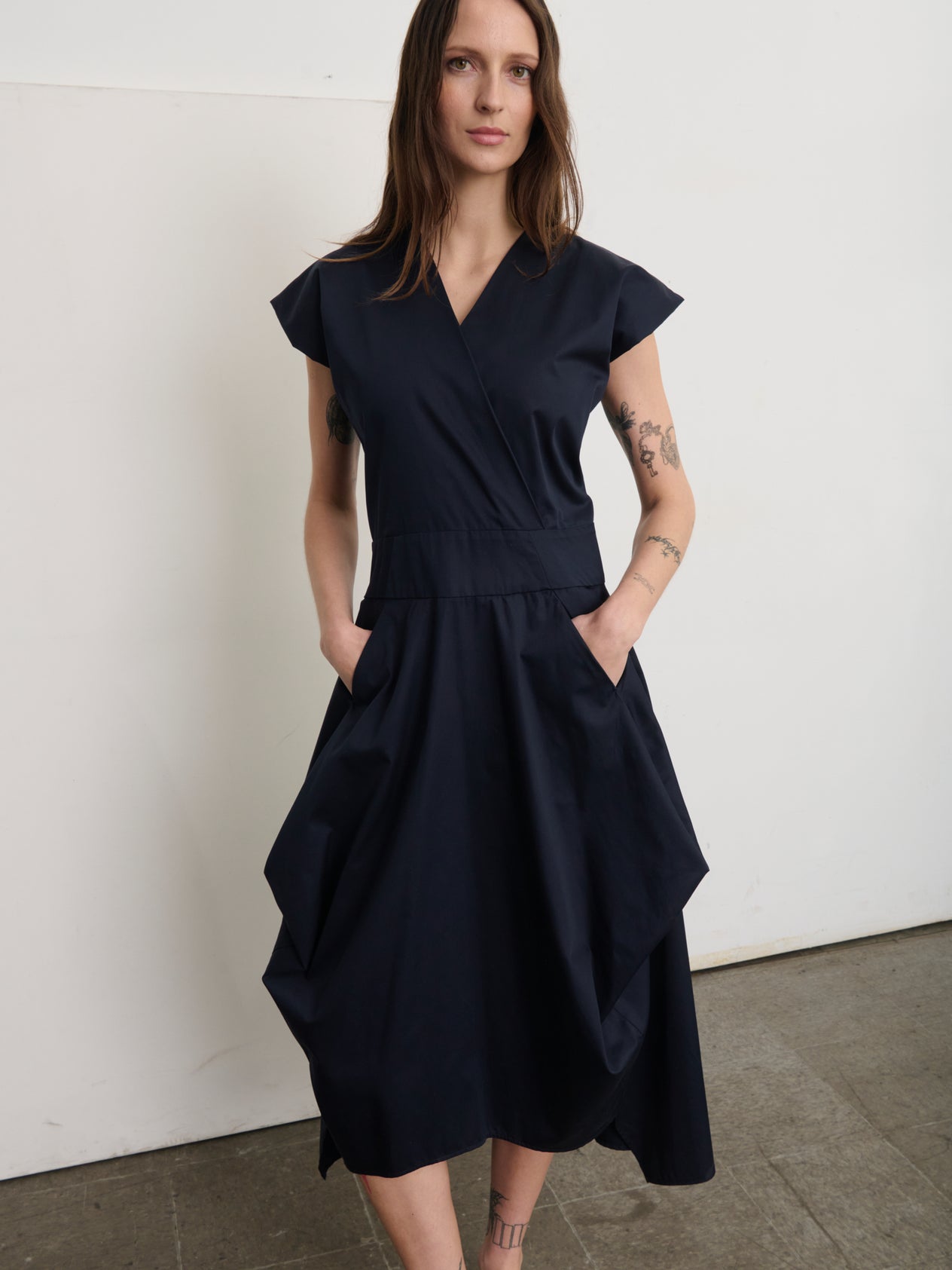 A person with long brown hair confidently wears a sleeveless, dark Wrap Nio Dress by Zero + Maria Cornejo. Tattoos adorn one arm, and they sport black slip-on shoes. Abstract shapes and a neutral wall in the background enhance the geometric charm.