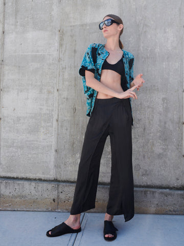 A person wearing oversized sunglasses, a blue patterned shirt, and a black crop top poses against a concrete wall. Their ensemble is elevated with Zero + Maria Cornejo's Ido Pant featuring a faux wrap overlay and paired with black slide sandals for an edgy yet chic look.