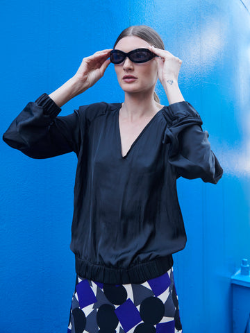 Against a bright blue background, a woman wears black oval sunglasses and the Cuadro B Top by Zero + Maria Cornejo. Her skirt is a wardrobe staple with a bold geometric pattern in black, white, and blue. She adjusts her sunglasses with both hands.