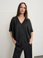 A person with long brown hair stands against a plain white background, wearing Zero + Maria Cornejo's Circle Top in black washed georgette with matching pants. The draped silhouette complements their neutral expression as they face the camera, hands in pockets.