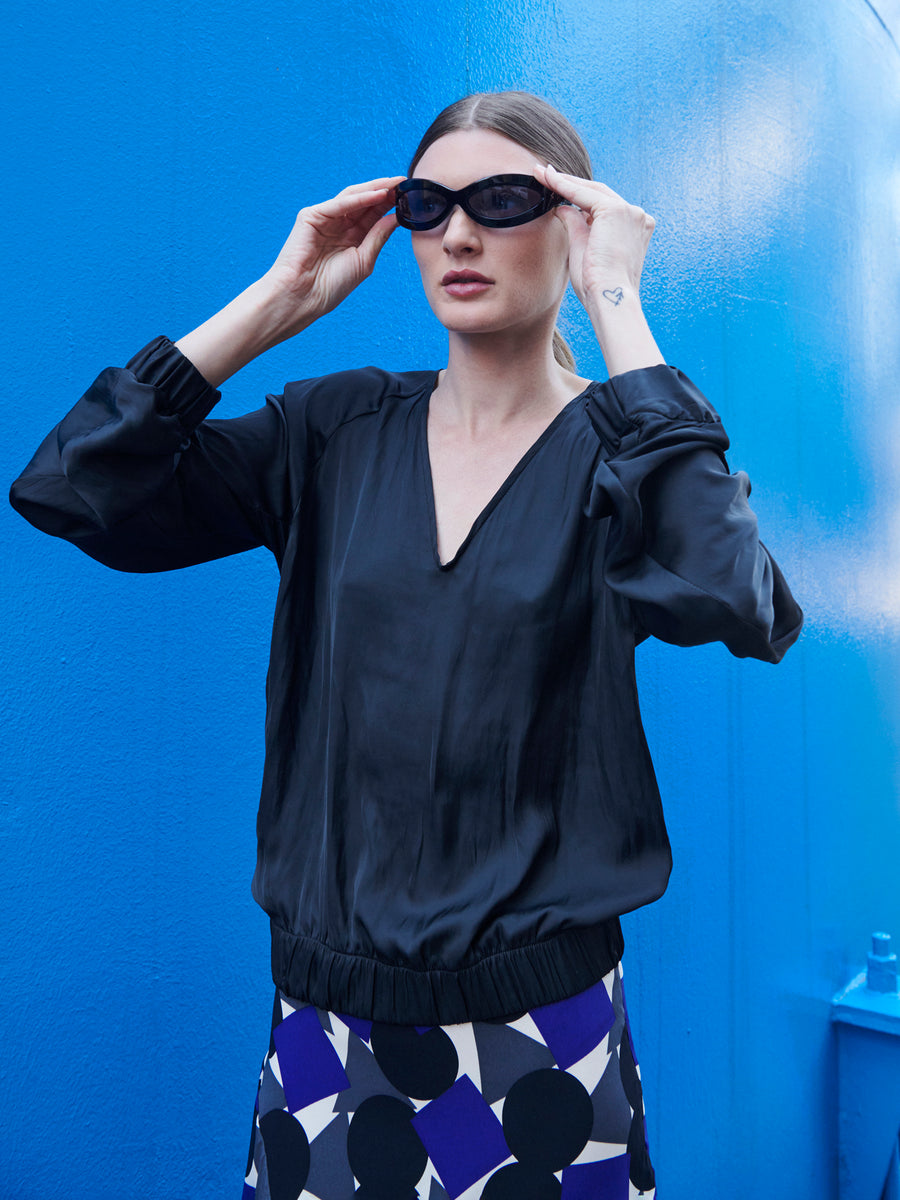 Against a bright blue background, a woman wears black oval sunglasses and the Cuadro B Top by Zero + Maria Cornejo. Her skirt is a wardrobe staple with a bold geometric pattern in black, white, and blue. She adjusts her sunglasses with both hands.