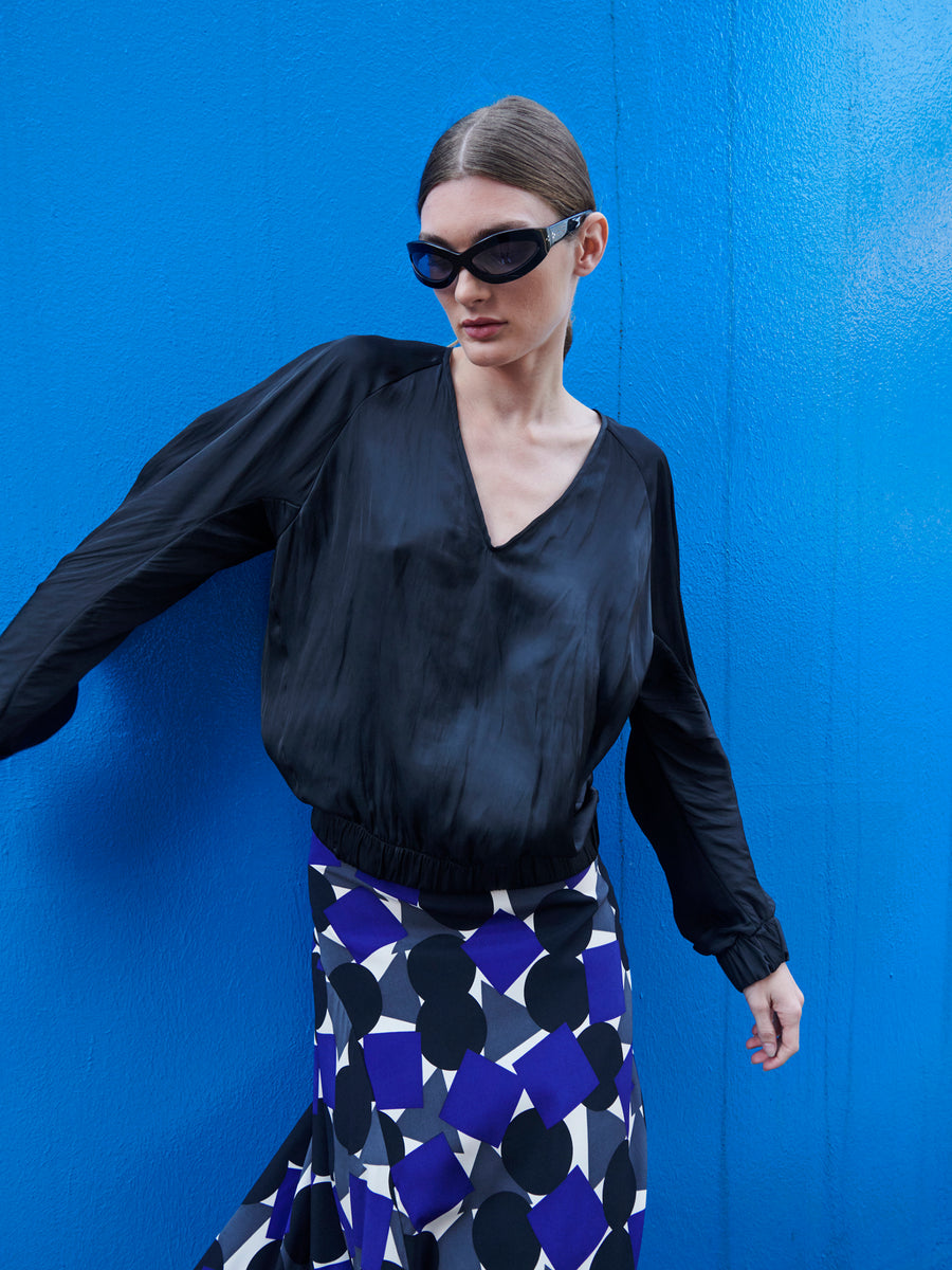 A person with sunglasses and slicked-back hair poses by a blue wall in a stylish Cuadro B Top from Zero + Maria Cornejo and a geometric-patterned skirt in black, white, and blue, highlighting the blend of modernity and chic versatility found in packable wardrobe staples.