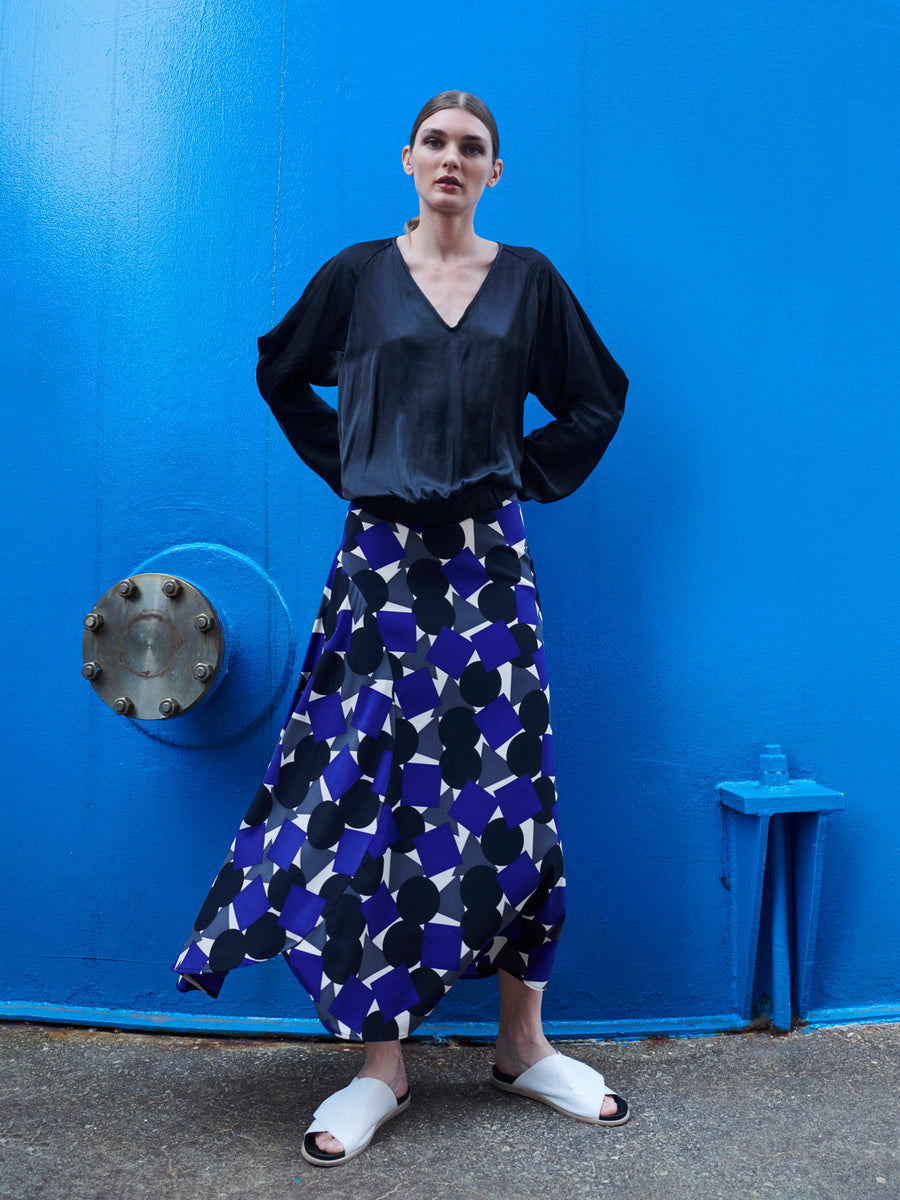 Against a bright blue wall, a person models the Cuadro B Top by Zero + Maria Cornejo and a flowing skirt with a striking geometric pattern in blue, white, and black. Paired with white slide sandals, they embody effortless chic confidence.